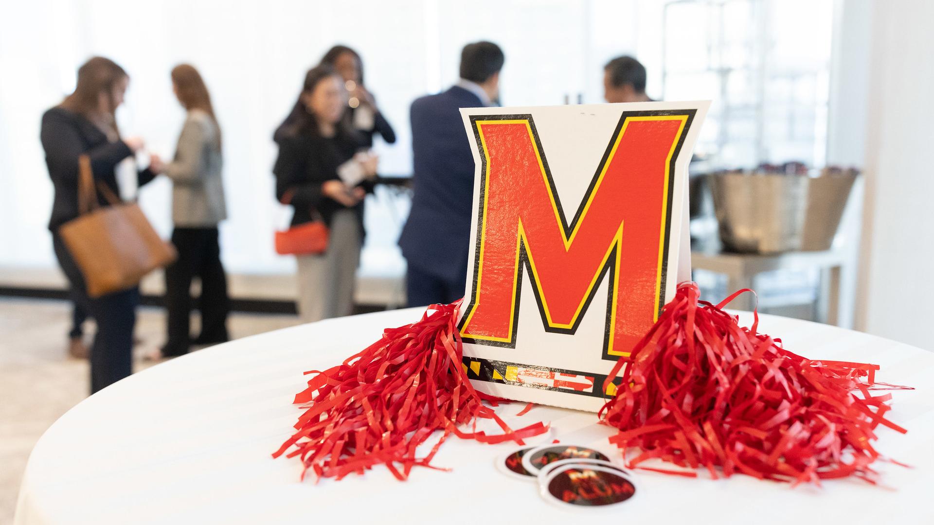 A Maryland "M" cardboard logo with pompoms in focus, and people networking in the background