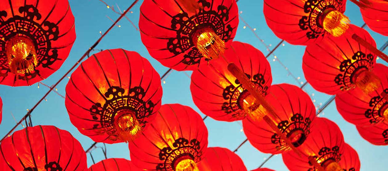 Red paper lanterns with gold decorative details hang overhead against a bright blue sky, displayed in rows as part of a Lunar New Year celebration.