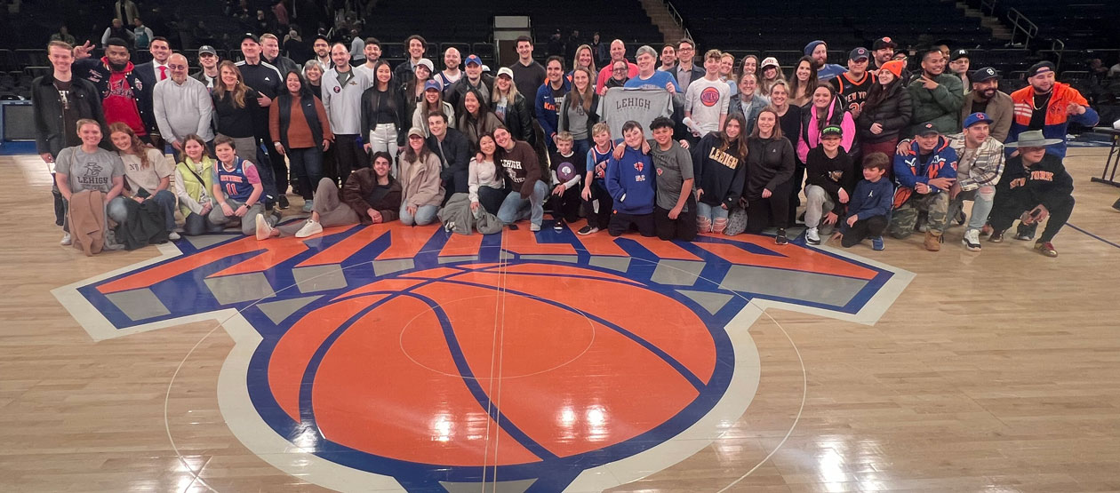 a large group of people gather on the Knicks' basketball court around their painted team emblem for a  photo