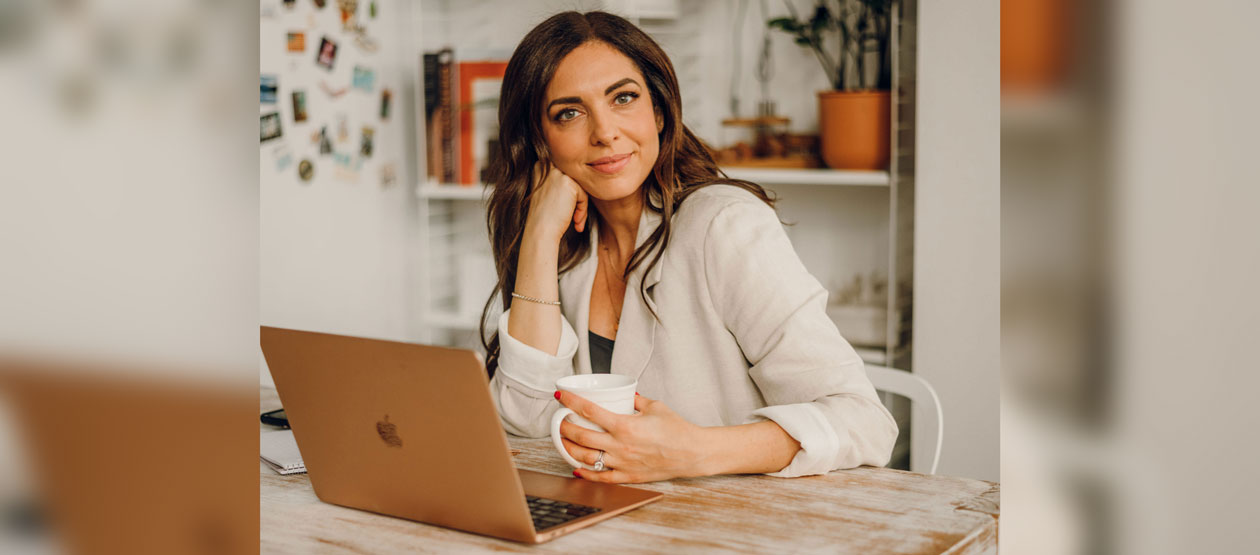 Maura Walters sits at a table, laptop in front of her and coffee mug in her hand, smiling at the camera with her head resting on her hand